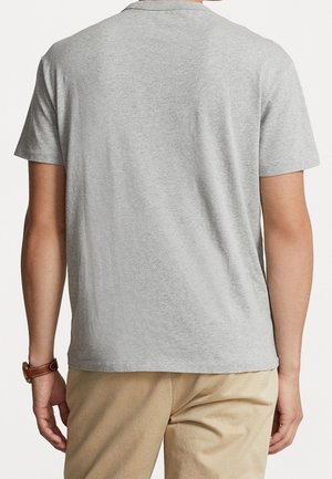 Mann, der ein schlichtes hellgraues kurzärmeliges T-Shirt und beige Hose trägt, von hinten gesehen, mit einer braunen Armbanduhr am linken Handgelenk.