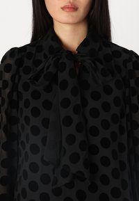 Blouse noire à pois avec un tissu transparent, dotée d'un col à nouer et de longues manches. Les pois sont prononcés et texturés.