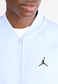 Svijetloplava zip-up jakna od glatkog materijala, s crnim izvezenim Jumpman logom na lijevoj strani grudi i rebrastim ovratnikom.