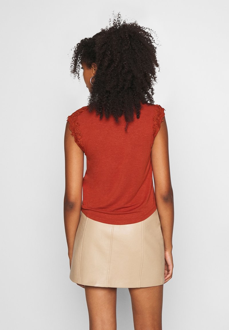 Femme aux cheveux bouclés portant un haut rouge sans manches avec des détails en dentelle et une mini-jupe en cuir beige, debout de dos.