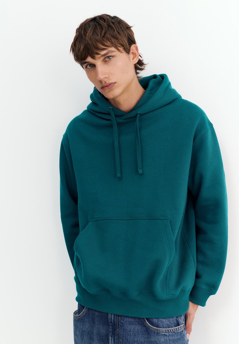 Jeune homme aux cheveux bruns portant un sweat à capuche bleu sarcelle et un jean bleu, debout devant un fond blanc, regardant légèrement sur le côté.