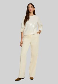 Pull crème à sequins avec manches larges, assorti à un pantalon en velours côtelé beige clair. La tenue est complétée par des bottines marron.