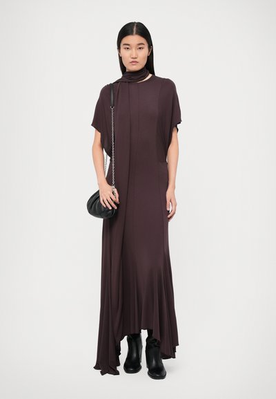 Long, sleeveless dress in deep purple fabric with a flowing design, featuring draped details and a high slit. Black handbag and knee-high boots.