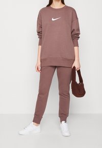 Sudadera marrón de Nike con un logo blanco, combinada con pantalones de chándal marrones a juego y zapatillas blancas. Sosteniendo un bolso texturizado marrón.