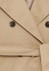 Beżowy płaszcz trench z szerokimi klapami, ciemnobrązowymi guzikami i pasującym paskiem z tkaniny związanym w talii.