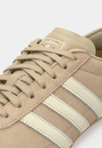 Μπεζ σνίκερ από σουέντ με τρεις διαγώνιες λευκές ρίγες, υφασμάτινο επάνω μέρος και ελαστική σόλα. Διαθέτει ανάγλυφο λογότυπο στη γλώσσα.