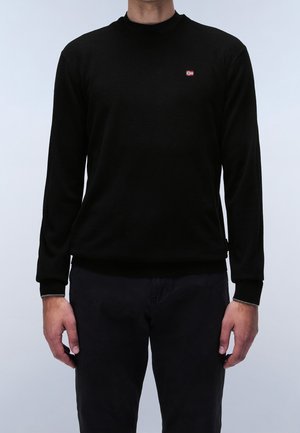 Mand iført en ensfarvet sort langærmet sweater med et lille rødt og hvidt flagemblem på venstre bryst, kombineret med sorte bukser.
