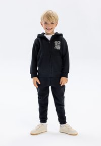 Schwarze Zip-Hoodie mit Fronttaschen und Logodetail, kombiniert mit passenden schwarzen Hosen. Das Modell trägt cremefarbene Sneaker und weiße Socken.