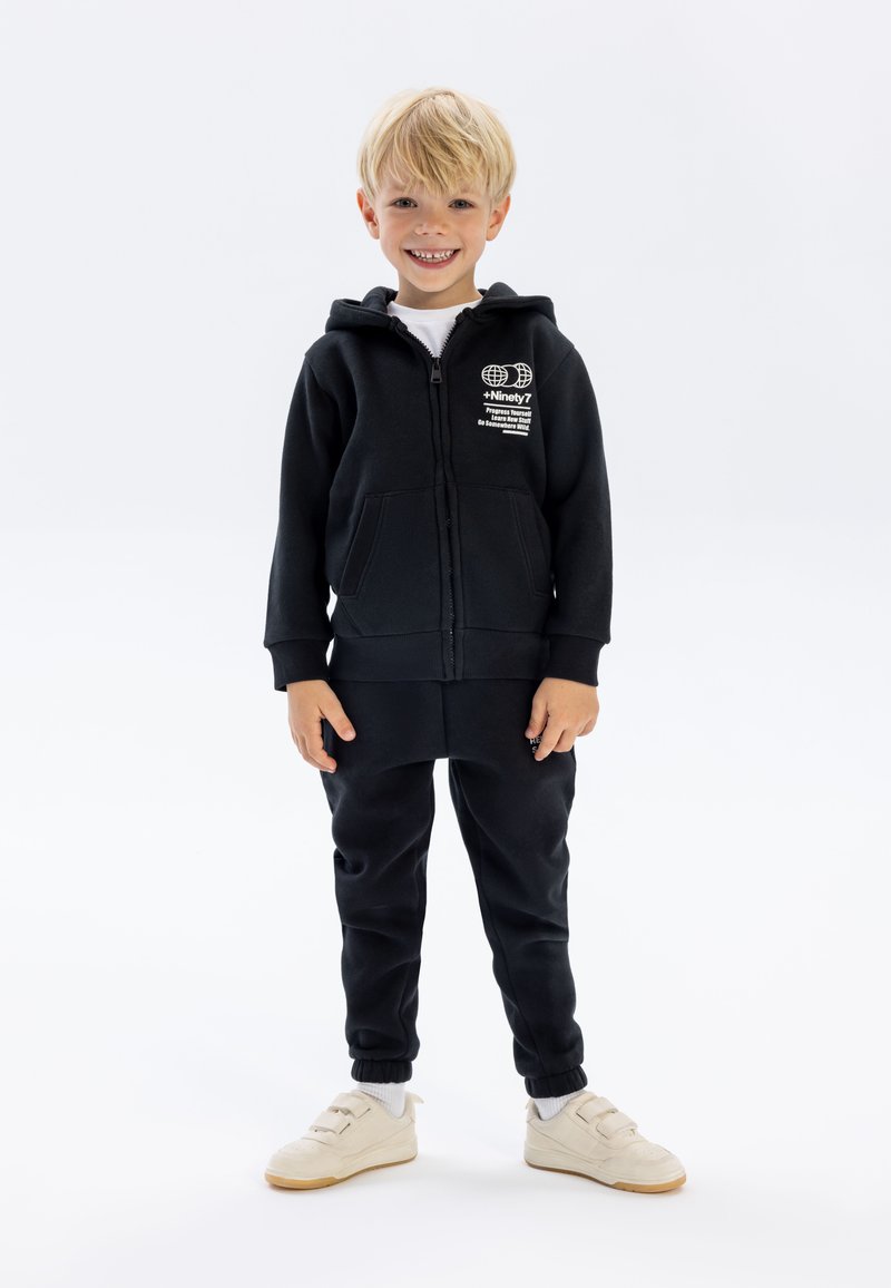 Schwarze Zip-Hoodie mit Fronttaschen und Logodetail, kombiniert mit passenden schwarzen Hosen. Das Modell trägt cremefarbene Sneaker und weiße Socken.