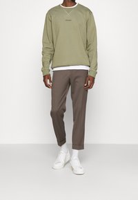 Uomo che indossa un felpa verde oliva con la scritta "LES DEUX", pantaloni marroni con risvolto, calze bianche e sneakers bianche, in piedi su uno sfondo neutro.