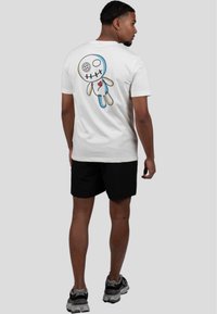 Camiseta blanca de manga corta con un colorido diseño de muñeca bordada en la parte trasera. Se lleva con pantalones cortos negros y zapatillas de correr grises.
