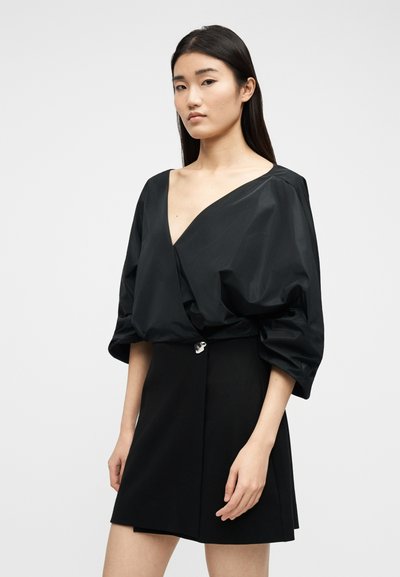 Femme portant une robe noire à décolleté en V profond et manches volumineuses, debout devant un fond gris clair uni.