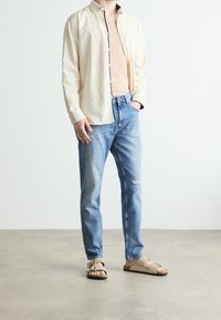 Beige långärmad skjorta, räfflad tan linne, ljusblå slitna jeans och beige sandaler med flera remmar och en enkel spänne.