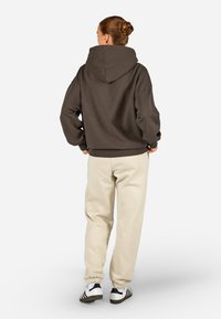 Brązowa bluza z kapturem z prążkowanymi mankietami i dołem, w połączeniu z kremowymi spodniami dresowymi oraz czarno-białymi butami sportowymi, widok od tyłu.