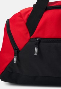 Rode en zwarte sporttas van textuurstof, met twee ritsen en Puma-branding. Compakt, hoekig ontwerp geschikt voor sportief gebruik.