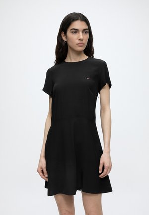 Pikkade tumedate juustega naine, kes kannab lühikeste varrukatega musta kleiti, millel on väike logo rinnal, seismas lihtsa heleda tausta ees.