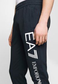 Černé tepláky s bílým grafickým logem na boku. Mají elastický pas a boční kapsy, vyrobené z měkké látky.