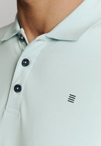 Lichtgroene polo met een textuurstof, twee donkere knopen en een klein zwart logo met drie horizontale lijnen op de borst.