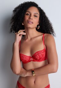 Soutien-gorge en dentelle rouge avec broderies florales, soutien-gorge à armatures, bretelles ajustables et bande satinée. Présente des détails logo en or.