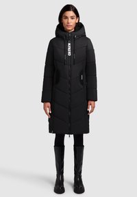 Manteau d'hiver noir long avec une capuche, matelassage en chevrons, fermeture éclair, poches latérales et cordons de serrage réglables. Porté avec des bottes noires jusqu'au genou.