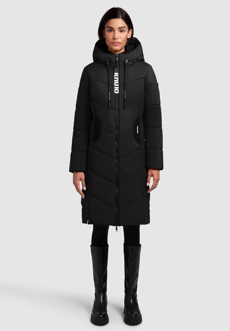 Manteau d'hiver noir long avec une capuche, matelassage en chevrons, fermeture éclair, poches latérales et cordons de serrage réglables. Porté avec des bottes noires jusqu'au genou.