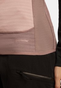 Primo piano di una persona che indossa una maglietta rosa Icebreaker e pantaloni neri con una tasca con zip, mano visibile di lato.
