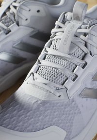 Lys grå atletiske sko med mesh-detaljer, syntetiske overlays og metalliske accenter. Har en struktureret overdel og flade snørebånd.
