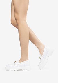 Mocassins blancs en cuir à enfiler avec une semelle blanche épaisse, présentant un accent en métal doré et un motif cousu sur le dessus.