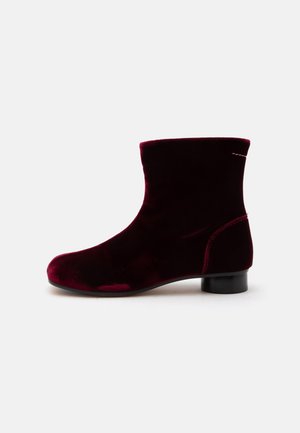Bota única de terciopelo hasta el tobillo en color burdeos oscuro con tacón bloque bajo negro y detalles mínimos de costura, mostrada de lado.