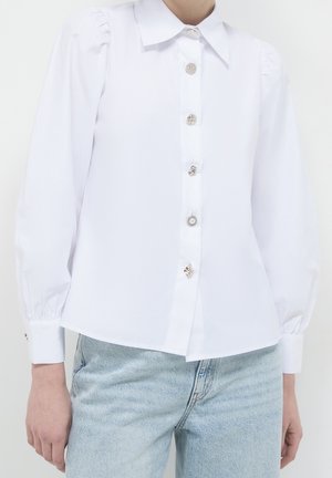 Blouse blanche à manches longues avec épaules bouffantes et boutons argentés décoratifs, portée avec un jean en denim bleu clair.