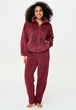 Lachende vrouw met krullend haar die een bijpassende zachte bordeauxrode fleecejas en broek draagt, blootsvoets staand tegen een witte achtergrond.