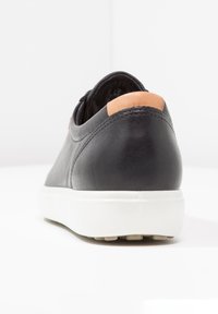 Zapatilla negra de cuero con suela blanca y lengüeta trasera en color beige, mostrada desde atrás sobre una superficie y fondo blancos.