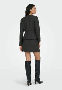 Šedý přiléhavý blazer a odpovídající sukně, doplněné černými koženými vysokými jehlovými botami. Ušité s důrazem na detaily, hladká textura a elegantní silueta.