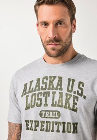 Skægget mand iført en lys grå skjorte med grøn tekst der står "ALASKA U.S. LOST LAKE TRAIL EXPEDITION," som viser en del af en tatoveret arm.