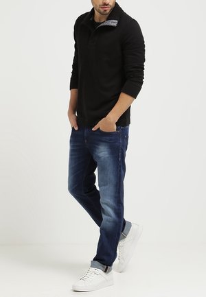 Sort langærmet polo-shirt med tekstureret krave, kombineret med mørkeblå jeans og hvide sneakers. Afslappet pasform med et enkelt design.