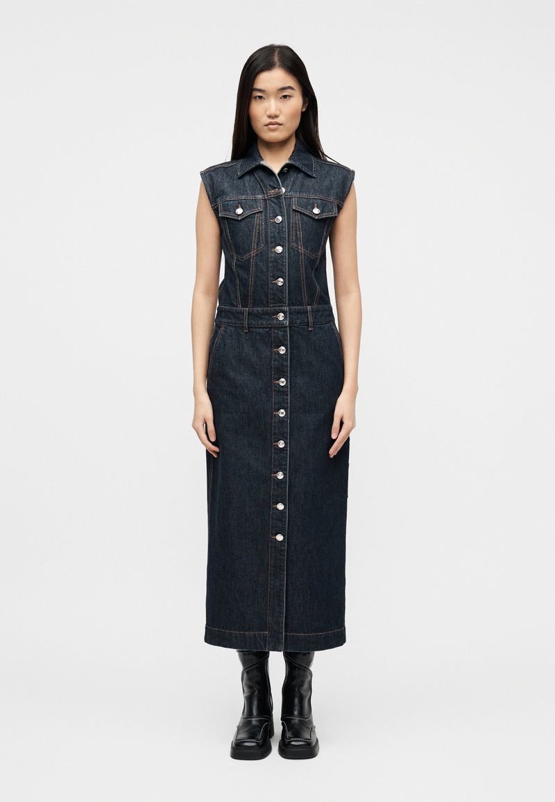 Γυναίκα που στέκεται φορώντας ένα  μαν sleeveless σκούρο τζιν φόρεμα με κουμπιά μπροστά και γιακά, μαζί με μαύρες μπότες μέχρι το μέσο της γάμπας, σε απλό φωτεινό φόντο.