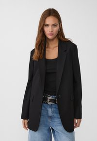 Schwarzer Blazer mit strukturiertem Design und einem einzigen Knopfverschluss, kombiniert mit einem schwarzen gerippten Tank-Top und einem genagelten Gürtel, getragen mit blauen Jeans.