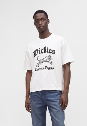 Мъж, носещ бяла тениска с графика на скок на тигър и текст "Dickies Campus Tigers", комбинирана със сини дънки.