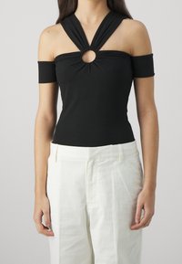 Top negro sin mangas con un detalle de corte circular en la parte delantera, hecho de una tela suave, combinado con pantalones blancos de corte a medida.