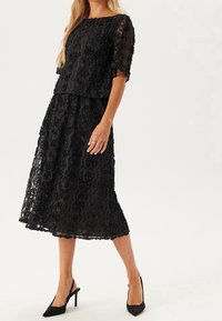 Schwarzes Spitzenkleid mit Blumenmuster, kurzen Ärmeln und einem knielangen Rock. Getragen mit schwarzen Stöckelschuhen. Glatte Textur.