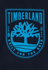 Un tissu bleu marine présente un logo Timberland turquoise, mettant en avant un design d'arbre avec le texte "CONÇU POUR LES AUDACIEUX" et "ÉTABLI EN 1973." Style graphique audacieux.
