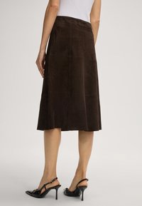 Bruine suède midi-rok met zijnaad en een gladde textuur. Gecombineerd met zwarte gehakte schoenen met een band over de enkel.
