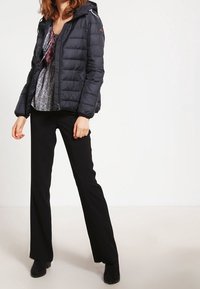 Námořnicky modrá prošívaná bunda s kapucí, předním zipem a bočními kapsami, nošená přes vzorované tričko a černé kalhoty.