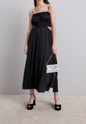 Kobieta ubrana w czarną, bezrękawnikową sukienkę midi z wycięciami po bokach, czarne buty na platformie oraz trzymająca małą srebrną torebkę z złotym łańcuszkiem.