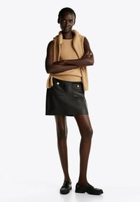 Beige kabelbrei sleeveless top gecombineerd met een zwarte leren mini rok met twee voorkoeken en gouden knoopaccenten. Zwarte loafers.