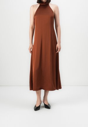 Femme portant une robe midi marron sans manches à col halter avec des chaussures noires à talons bas, debout devant un fond blanc uni.