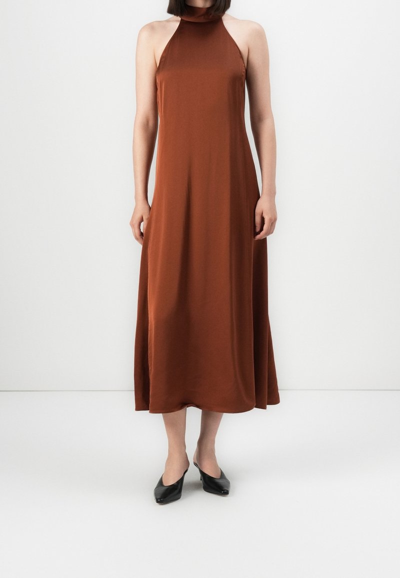 Femme portant une robe midi marron sans manches à col halter avec des chaussures noires à talons bas, debout devant un fond blanc uni.