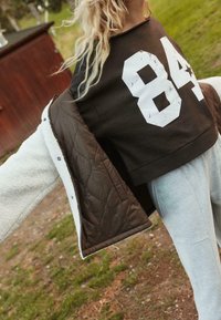 Giacca trapuntata marrone con esterno in pile bianco, indossata sopra una felpa nera con una grande stampa bianca "84". Pantaloni della tuta grigi visibili.