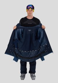 Mørkeblå skijakke holdt åben, som viser et glat indre foret, struktureret design, justerbare elementer og lynlåslommer. Snedérk støvler på.