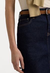 Jupe en denim foncé avec des poches visibles et des détails de couture. Une ceinture en cuir marron souligne la taille. Texture lisse, design ajusté.